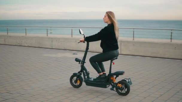 Mujer rubia bonita conduciendo bicicleta eléctrica cerca del mar . — Vídeos de Stock