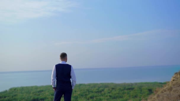 Ung brudgum står nära kullen kusten med havet på bakgrunden — Stockvideo