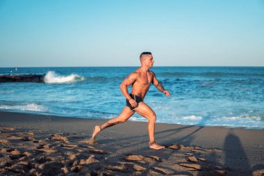 Tattooed bodybuilder sexy male coach at the beach. clipart