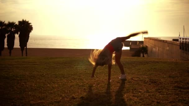 Boldog lány ugrik, és ezzel cartwheels a strandon élvező naplemente — Stock videók
