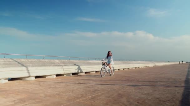 Modelo asiático en un vestido azul monta una bicicleta blanca en la terraza del mar — Vídeos de Stock