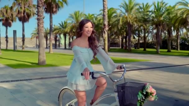 Modelo asiático en un vestido azul monta una bicicleta blanca en un parque verde — Vídeos de Stock
