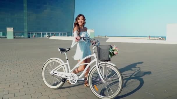 Modelo asiático en un vestido azul monta una bicicleta blanca en la terraza del mar — Vídeo de stock