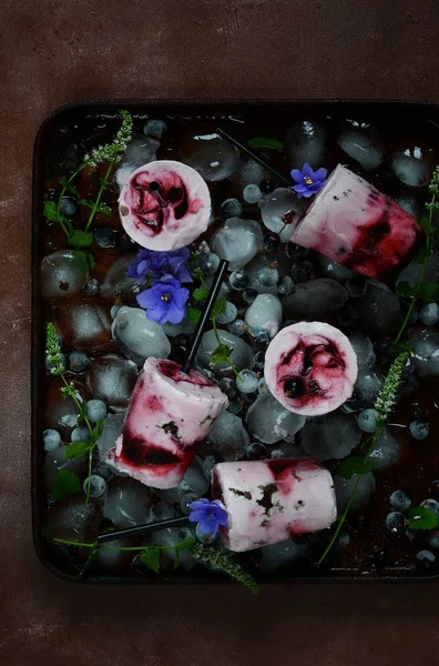 Kremalı dondurma siyah frenk üzümü, böğürtlen, ahududu, nane ve menekşe koyu arka plan üzerinde çiçekler meyveler. Yaz soğutma tatlı. Ev yapımı dondurma veya çubuklu dondurmayı yeşil nane bırak dekore edilmiş