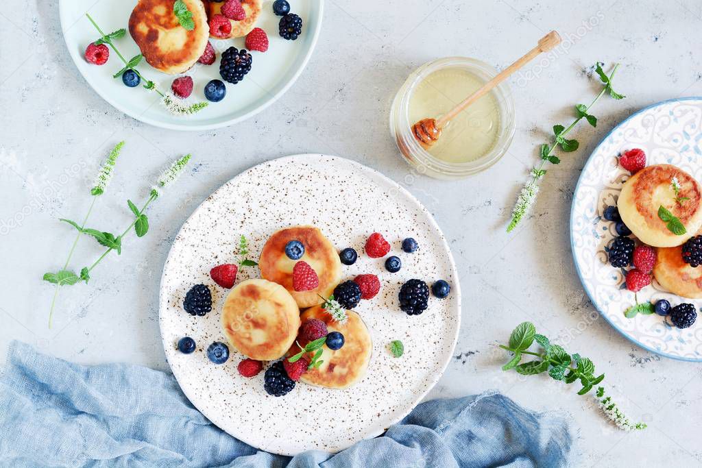  Sunday breakfast with cheesecake, honey, fresh berries  and mint. Cottage cheese pancakes or curd fritters decorated honey and berries in plate on blue table top view. Healthy and diet breakfast.