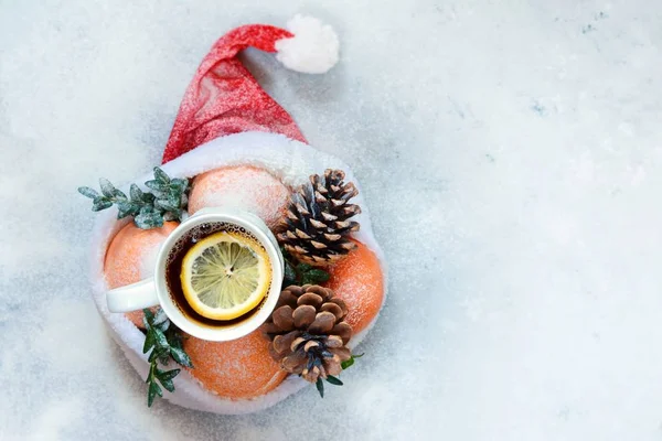 Ripe oranges, cup of tea with lemon, cones in the hat of Santa Claus with boxwood plant on a blue background. Festive mood, Christmas and New Year. Copy space