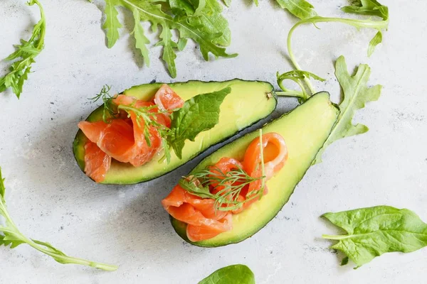 Ripe avocado boats with slices of salted trout, salmon and fresh greens on a gray background. Avocado boats stuffed with salmon with lime, spinach leaves and arugula, concept healthy food, diet.