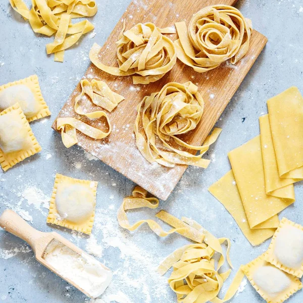 Ev yapımı İtalyan makarnası, ravioli, fetuçin, ahşap tahtada tagliatelle ve mavi arka planda. Pişirme süreci, çiğ makarna. Lezzetli çiğ ravioli, ricotta ve ıspanak, arka planda un.