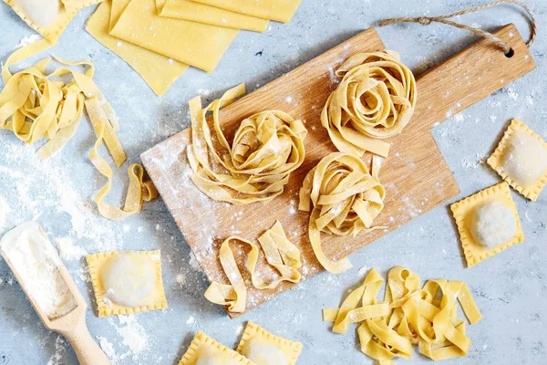 Ev yapımı İtalyan makarnası, ravioli, fetuçin, ahşap tahtada tagliatelle ve mavi arka planda. Pişirme süreci, çiğ makarna. Lezzetli çiğ ravioli, ricotta ve ıspanak, arka planda un.