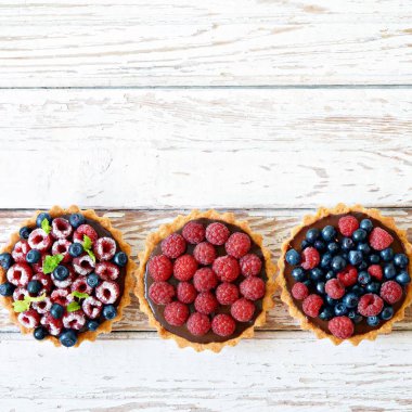 Çikolata ganache 'li ahududu ve yaban mersinli tartlar, taze böğürtlen ve nane yaprağı, seçici odaklanma. Beyaz arka planda taze meyveli tart, masada taze ev yapımı meyveli kek.