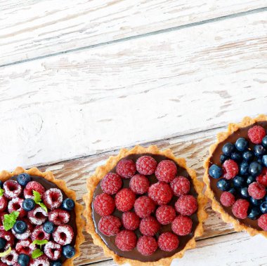 Çikolata ganache 'li ahududu ve yaban mersinli tartlar, taze böğürtlen ve nane yaprağı, seçici odaklanma. Beyaz arka planda taze meyveli tart, masada taze ev yapımı meyveli kek.