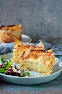 Patates graten filo hamur pişmiş, gevrek peynir kabuğu ve arugula bir salata ile, güneşte kurutulmuş domates ve mavi bir arka plan üzerinde peynir. Fransız mutfağından bir yemek