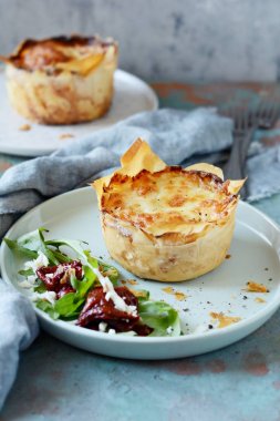 Patates graten filo hamur pişmiş, gevrek peynir kabuğu ve arugula bir salata ile, güneşte kurutulmuş domates ve mavi bir arka plan üzerinde peynir. Fransız mutfağından bir yemek