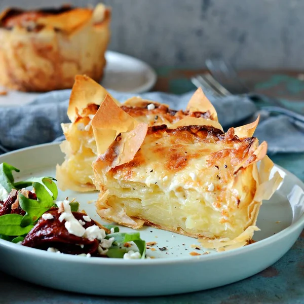 Patates graten filo hamur pişmiş, gevrek peynir kabuğu ve arugula bir salata ile, güneşte kurutulmuş domates ve mavi bir arka plan üzerinde peynir. Fransız mutfağından bir yemek