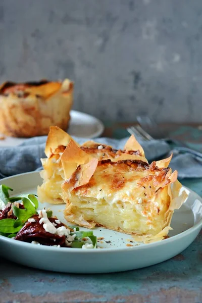 Patates graten filo hamur pişmiş, gevrek peynir kabuğu ve arugula bir salata ile, güneşte kurutulmuş domates ve mavi bir arka plan üzerinde peynir. Fransız mutfağından bir yemek