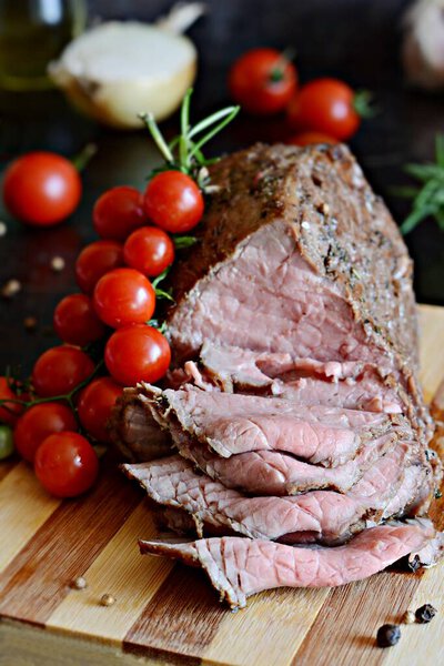 Slice of baked beef in spices on a wooden board with a sprig of cherry tomatoes and fresh rosemary. Juicy Barbecue Meat (roast beef) .