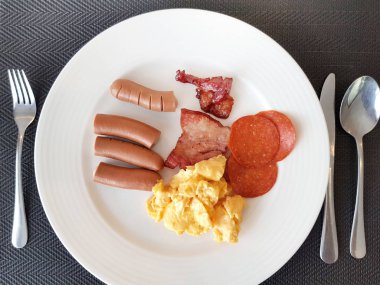 Otel kahvaltısı büfesi sabah yemeği, kızarmış domuz eti, sosis, jambon ve çırpılmış yumurta kaşıklı ve çatallı beyaz tabak..