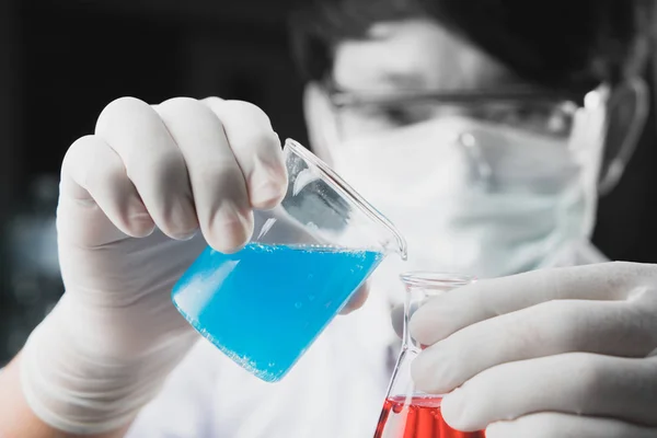 化学の学生の少年は 生化学実験で赤い液体でエルレマイアーフラスコ化学にビーガラスに青い液体を注ぐ 研究室で水を混ぜる — ストック写真