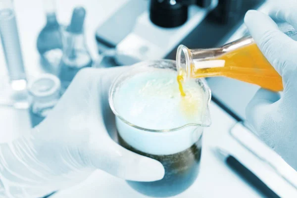 化学者の手を閉じて 生化学実験で青色の液体でエルレマイヤフラスコ化学にビールガラスに黄色の液体油を注ぐ 研究室で水を混ぜる — ストック写真