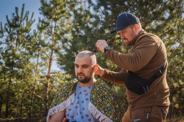 Tough red bearded guy shaving bold the other man outdoors in the pine forest
