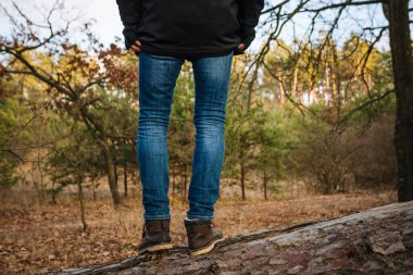 Mavi kotlu, kahverengi deri çizmeli, ağaç gövdesinde duran genç yürüyüşçü.