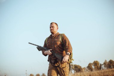 Kızıl sakallı güçlü genç avcı silahını tutuyor ve mavi gökyüzünün altında toprak yolda yürüyor.