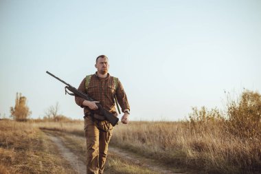 Kızıl sakallı güçlü genç avcı silahını tutuyor ve mavi gökyüzünün altında toprak yolda yürüyor.