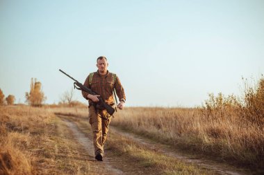 Kızıl sakallı güçlü genç avcı silahını tutuyor ve mavi gökyüzünün altında toprak yolda yürüyor.