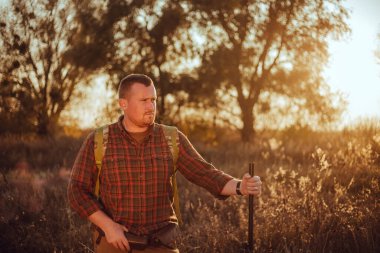 İrlandalı görünümlü kareli gömlekli kızıl sakallı avcı gün batımında dışarıda duruyor ve silahını tutuyor.