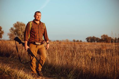Kızıl sakallı güçlü genç avcı silahını tutuyor ve mavi gökyüzünün altında toprak yolda yürüyor.