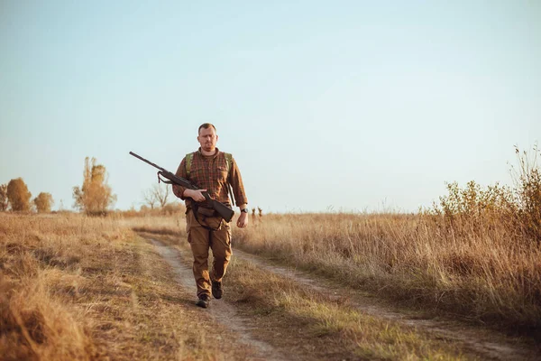 Kızıl sakallı güçlü genç avcı silahını tutuyor ve mavi gökyüzünün altında toprak yolda yürüyor.