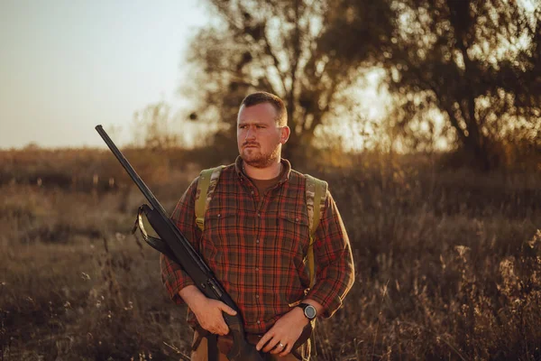 Irish looking red bearded hunter in checkered shirt staying outdoors in the sunset light and holding his gun