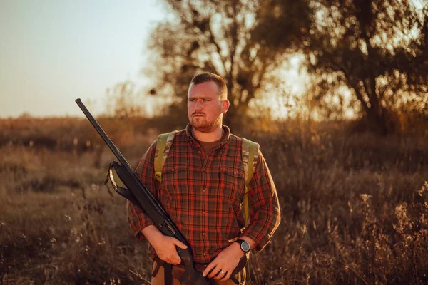 İrlandalı görünümlü kareli gömlekli kızıl sakallı avcı gün batımında dışarıda duruyor ve silahını tutuyor.