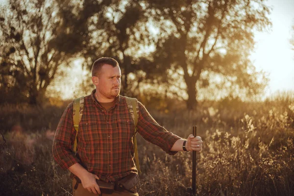 İrlandalı görünümlü kareli gömlekli kızıl sakallı avcı gün batımında dışarıda duruyor ve silahını tutuyor.
