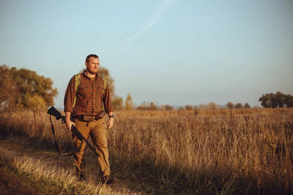 Kızıl sakallı güçlü genç avcı silahını tutuyor ve mavi gökyüzünün altında toprak yolda yürüyor.