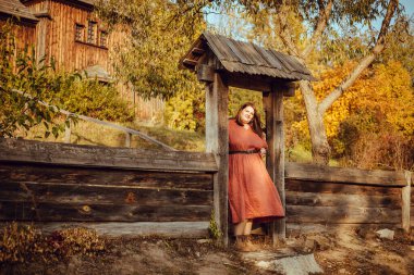 Güzel, kendine güvenen, kırmızı elbiseli ve kahverengi çizmeli bir model sonbahar manzarasında geleneksel Rus mimarisi ile dışarıda poz veriyor. 