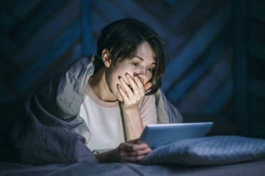 Evde korku filmi izleyen genç bir kadının portresi. Elleriyle ağzını kapatıyor ve bilgisayara bakıyor.