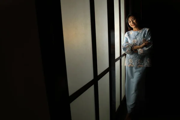 young Asian Malay woman wearing baju kurung dress at home in front window mood light on face dark background peaceful content calm tranquil break trough wonder