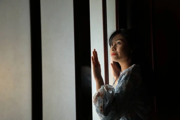 young Asian Malay woman wearing baju kurung dress at home in front window mood light on face dark background peaceful content calm tranquil break trough wonder