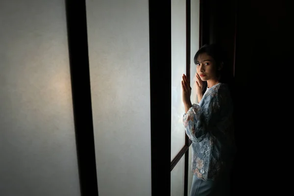 若いですアジアマレー人女性身に着けていますBaju Kurungドレスで自宅でフロントウィンドウ気分光上の顔暗い背景平和なコンテンツ穏やかな撮影静かな — ストック写真
