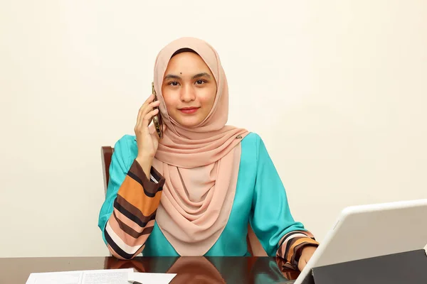 young Asian Malay Muslim woman wearing headscarf at home office student sitting at table talk mingle look at computer pone book document study discuss talk smile happy look at camera