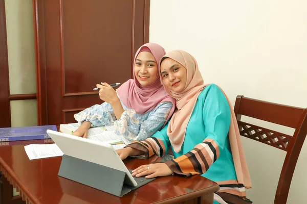 两名年轻的亚洲马来族穆斯林妇女头戴头巾坐在办公室里 坐在桌旁聊天看电脑书 讨论笑得开心 — 图库照片