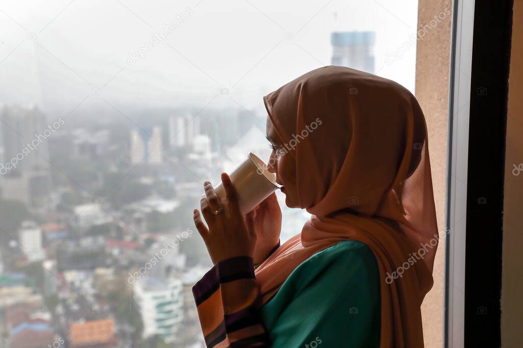 young Asian Malay Muslim woman wearing headscarf at home office student at window holding drinking coffee tea cup cityscape at background