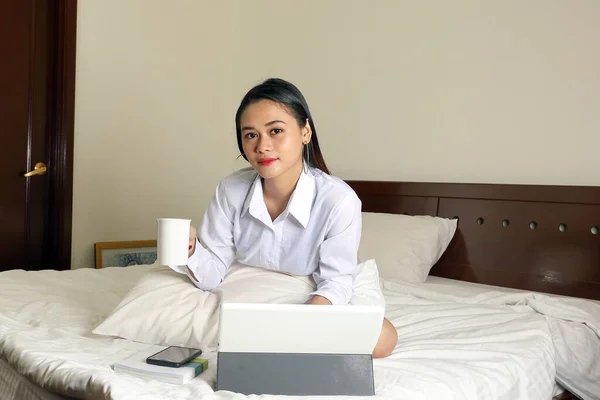 Young beautiful modern Malay woman sit on bed work study read write in bedroom laptop tab book smile look at camera