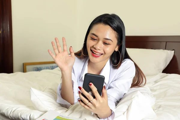 Young beautiful modern Malay woman sit on bed work study read write in bedroom laptop tab book smile look at camera