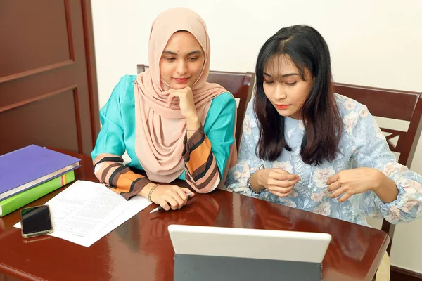 Two Young Asian Malay Muslim Woman Wearing Headscarf At Home Office Student Sitting At Table Talk Mingle Look At Computer Book Document Study Discuss Read Type Write Smile Happy Arab Pose