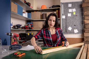 Bir cetvel ile çalışan ve kumaş atölyesinde masada ahşap üzerinde işaretleri yapma çalışkan odaklı profesyonel ciddi marangoz kadın yakından görünümü.