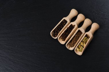 Tahta kaşıklar, tohum ve otlar karanlık taş tablosundaki çeşitli Hint baharat. Renkli baharat Kakuleden, kimyon, yenibahar, karabiber, üstten görünüm. Organik gıda, sağlıklı bir yaşam tarzı. Metin için yer