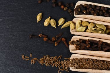 Tahta kaşıklar, tohum ve otlar karanlık taş tablosundaki çeşitli Hint baharat. Renkli baharat Kakuleden, kimyon, yenibahar, karabiber, üstten görünüm. Organik gıda, sağlıklı bir yaşam tarzı. Metin için yer