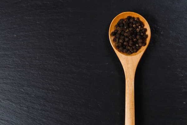 Black pepper spice in wooden spoon. Flat lay of black pepper spice on black stone. Black pepper spice, top view. Organic food, healthy lifestyle. Space for text